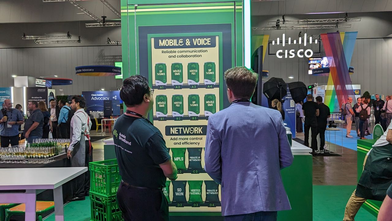 Aussie Broadband convenience store: Aussie Broadband's Cisco Live 2023 booth: A trade show booth resembling a convenience store. Two people, one wearing an Aussie Broadband shirt, arew staring at a sign meant to resemble a vending machine. Aussie Broadband Enterprise Technology offerings are listed on graphics of cans on the sign. "Network, add more control and efficiency", and "Mobile and Voice: reliable communication and collaboration".