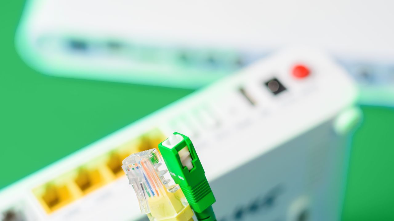 A photo of the ends of both a copper ethernet cable (on the left in yellow) and a fibre optic cable (on the right). In the background there is the back side of a white router/modem situated on a green surface.