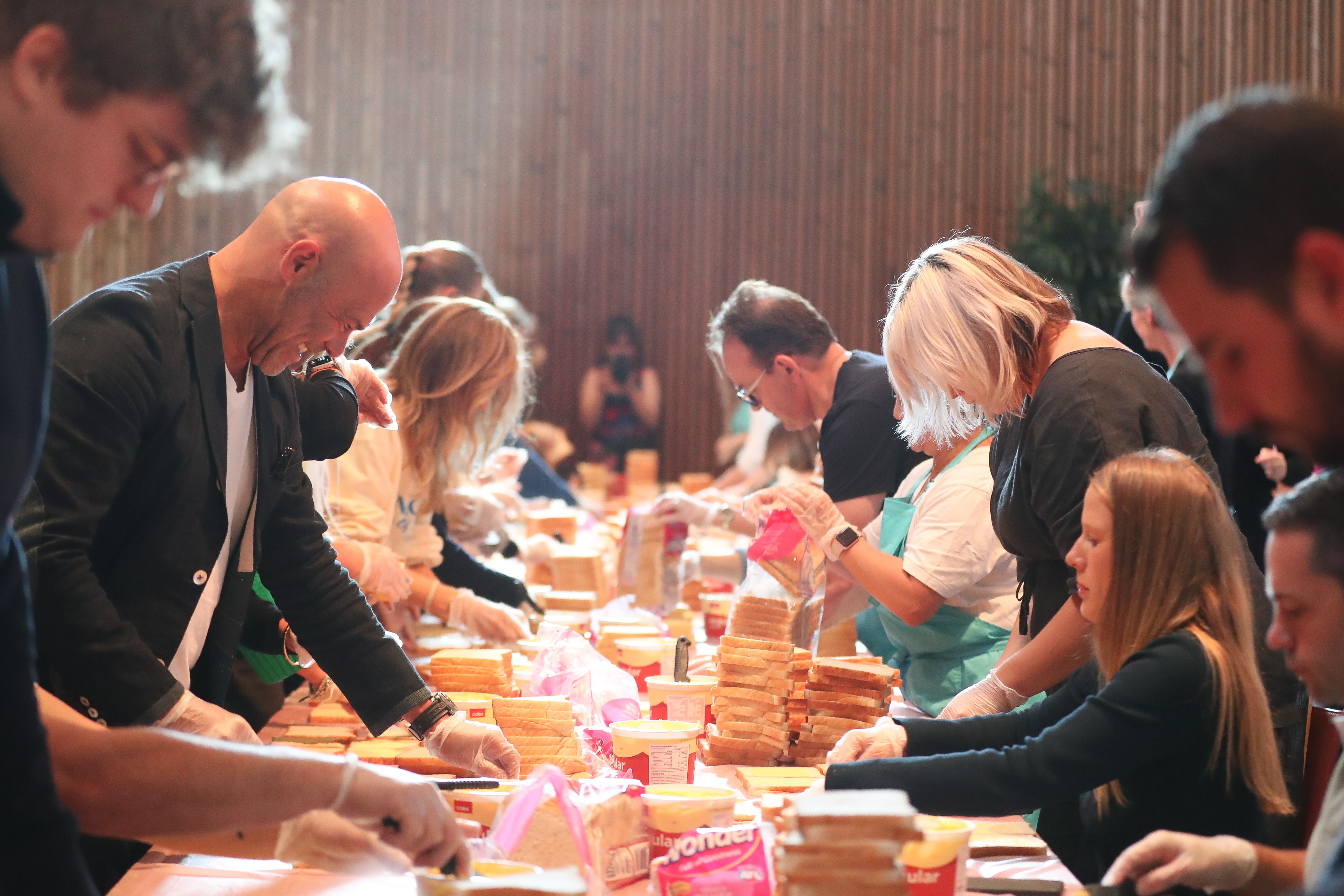 A row of people with many loaves of bread on front of them on a table, seemingly making sandwiches. There is a mix of different people, and some are seated whilst others are standing.