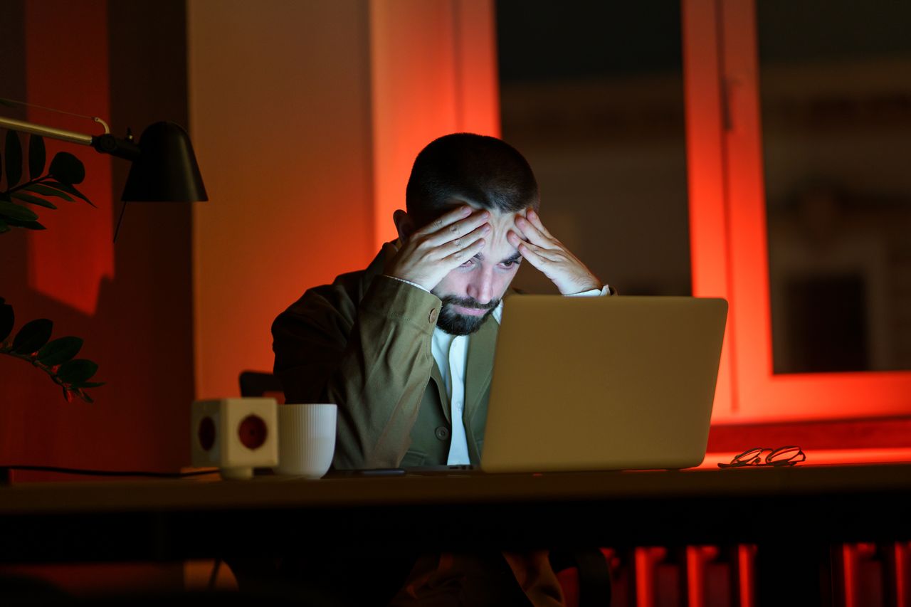 A photo of a man in a dark room, staring at his open laptop with his hands on his forehead, signifying that he is frustrated or confused by something.