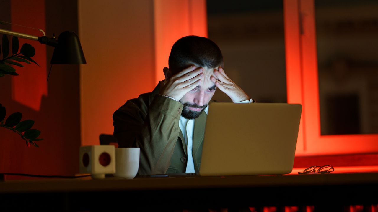 A photo of a man in a dark room, staring at his open laptop with his hands on his forehead, signifying that he is frustrated or confused by something.
