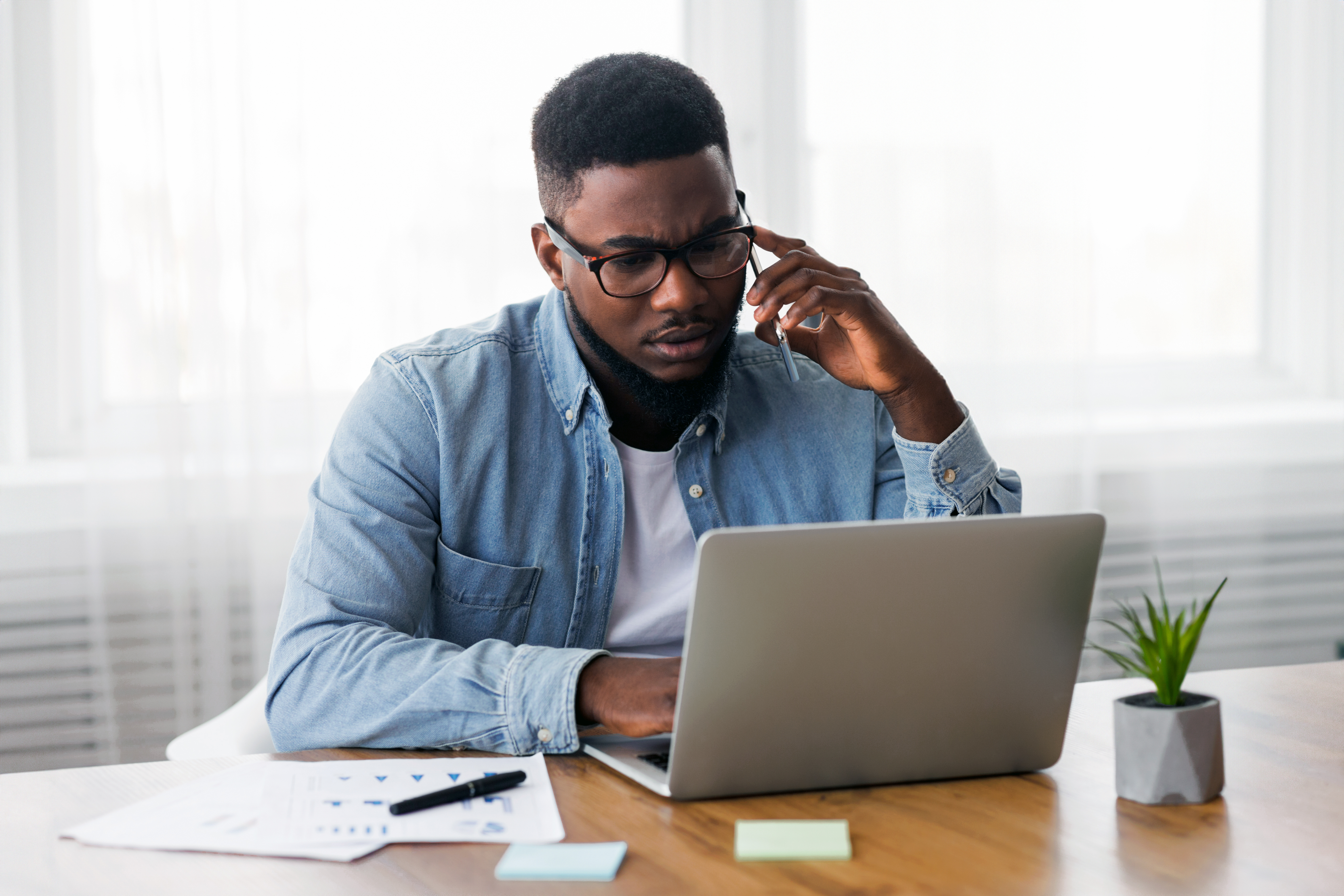 A photo of a gentleman on the phone, looking concerned, sitting at his laptop with papers beside him.