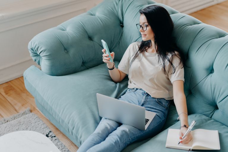 A photo of a female sitting on a blue, plushy couch with a phone in one hand, a pen in the other writing in a notebook, as well as a laptop on her lap.