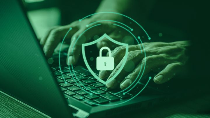 Hands typing on a laptop, which is placed on a desk. Superimposed onto the image is a slightly transparent green background, which gives the image a green tint. Superimposed at the front of the image is a locked padlock icon inside a shield. surrounding the shield are several circular lines with dots on each end.