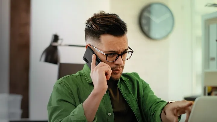 A man working from home pointing at a laptop screen while talking on a mobile phone