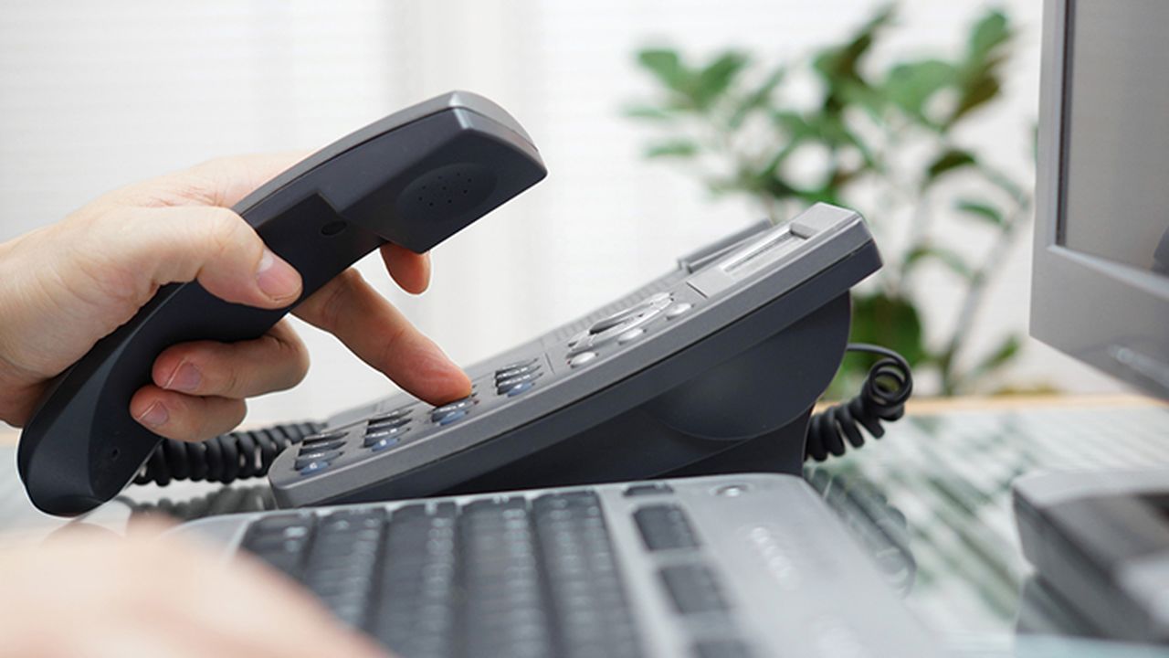 A phone handset placed on a desk. In front of it is a computer keyboard and screen. Visible are two hands, one hand is typing on the computer keyboard and the other is holding the phone while typing on the dial pad.