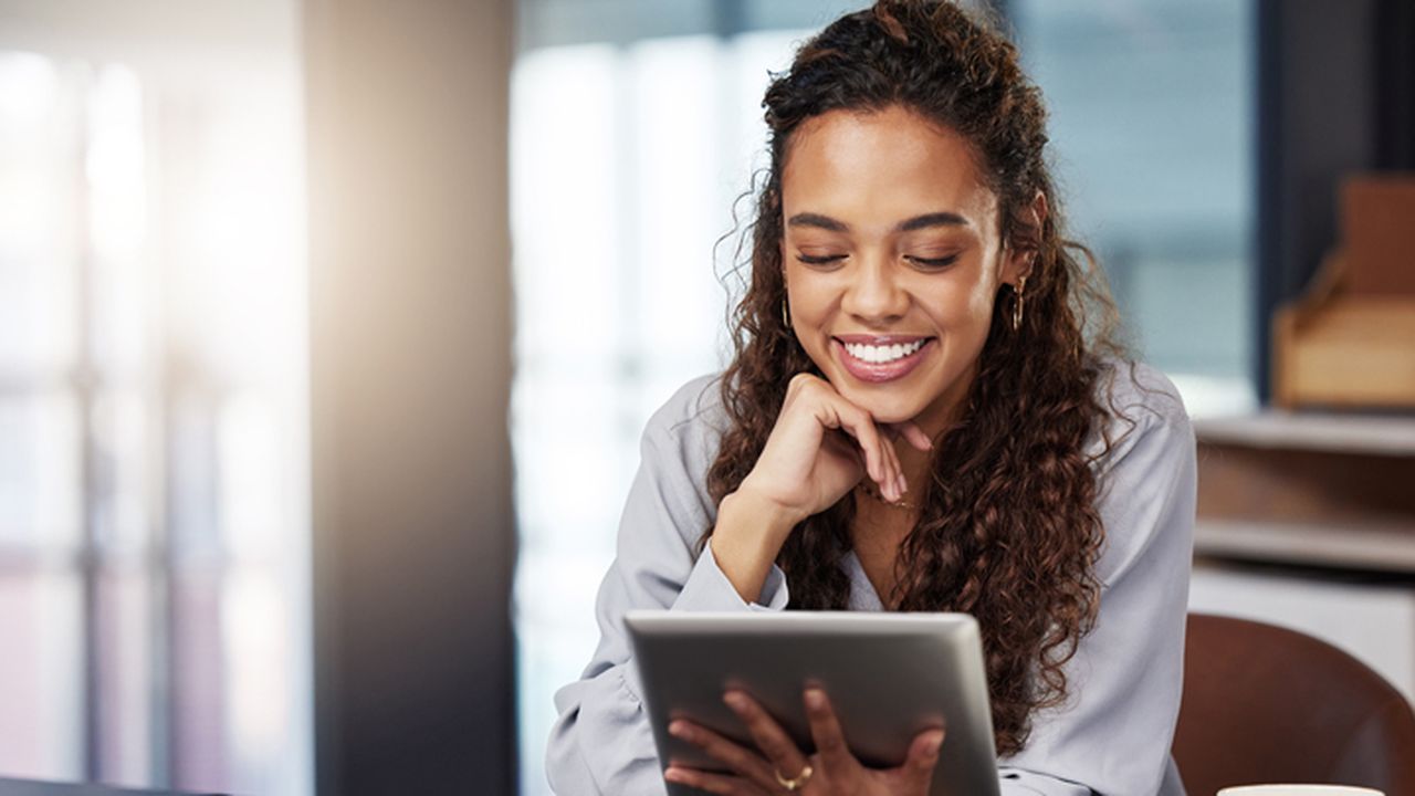A woman sitting at a desk in an office holding a tablet. She is looking at the tablet screen and smiling.