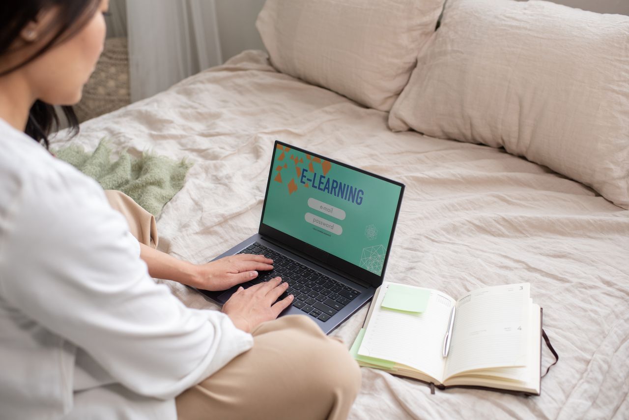 A lady sitting cross-legged on her bed, using her open laptop with "E-Learning" displayed on it. There is an open notebook to the right of the laptop, with a pen in it's spine