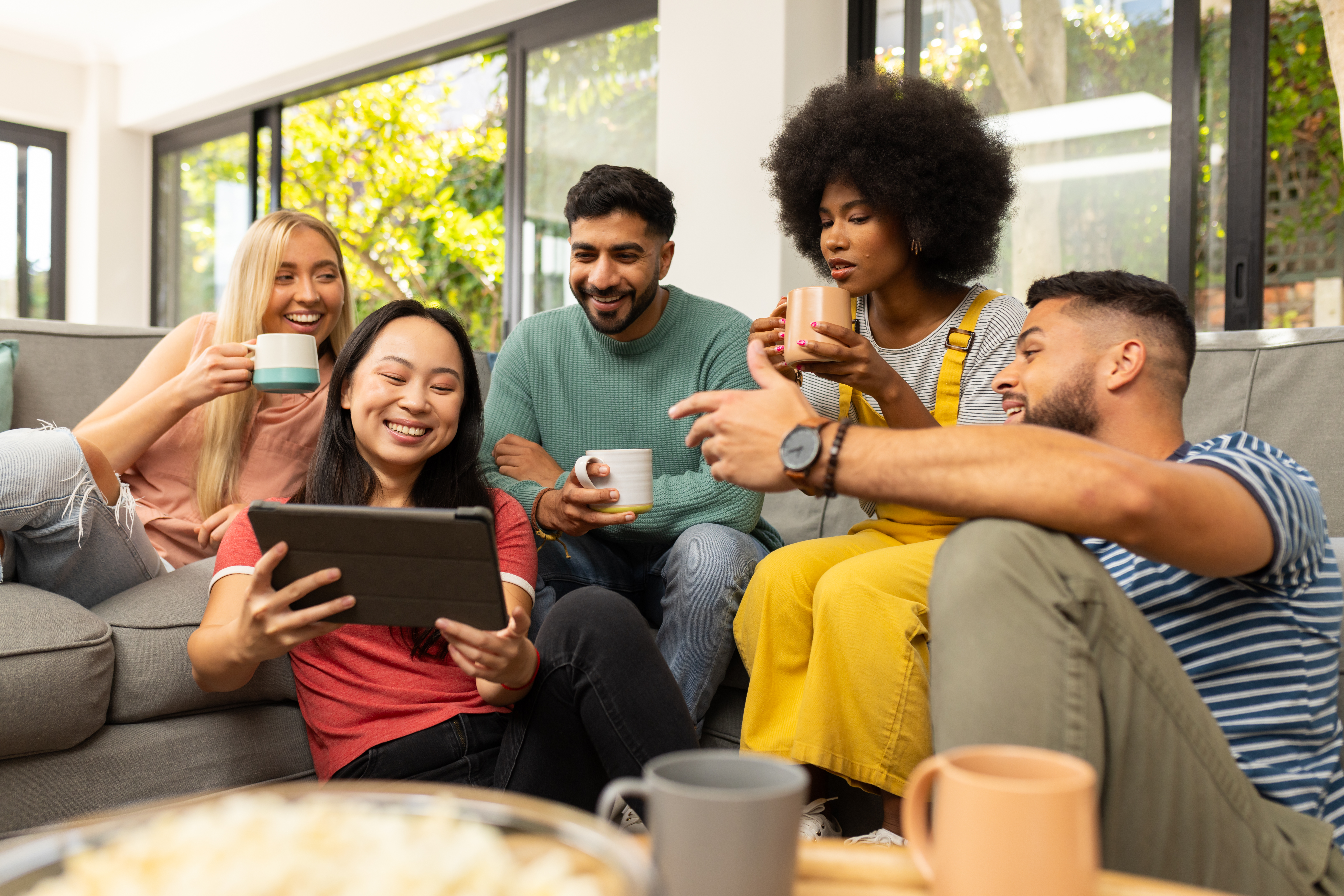 A group of 5 friends, sitting on a sofa drinking mugs of beverage while huddled around a tablet held by one of them.