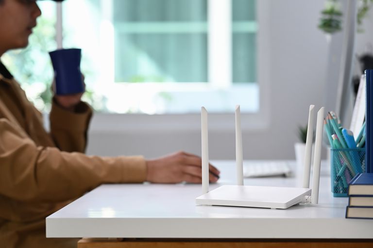 A photo of a white Wi-Fi router with 4 antennae, sitting on a white desk top. In the background, there is a person in a brown jacket sitting down, with a mug in one hand and a computer mouse in the other. Behind him, there is a window which shows a sunny outside environment.
