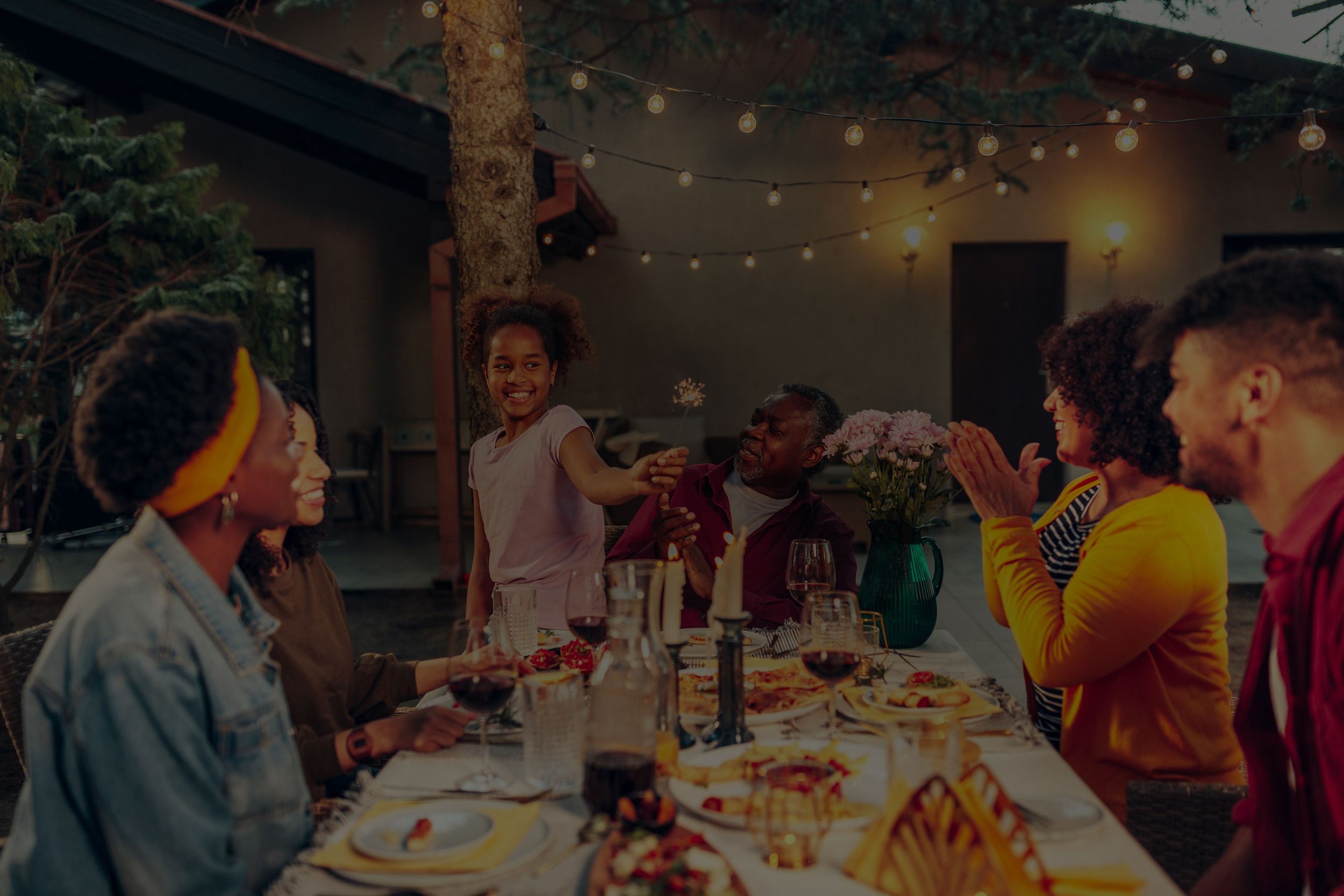 Family smiling on a backyard patio lit by electric string lights as a child holds a sparkler