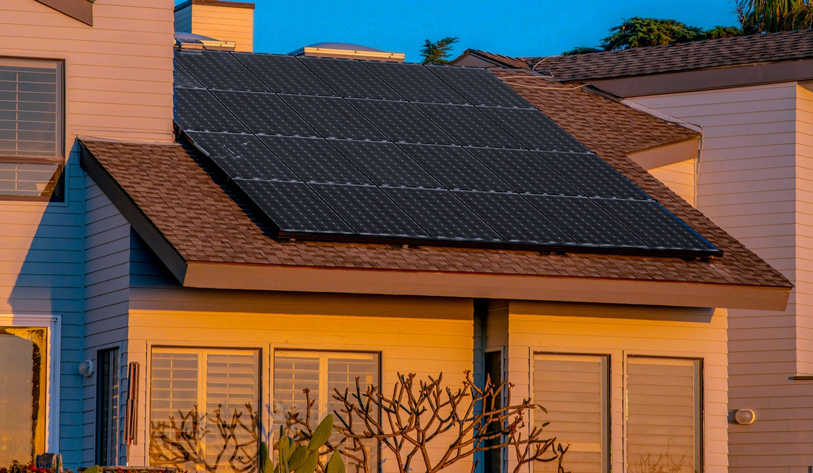 Rooftop solar panels on a modern house bask in golden sunset light, surrounded by windows and a few plants.