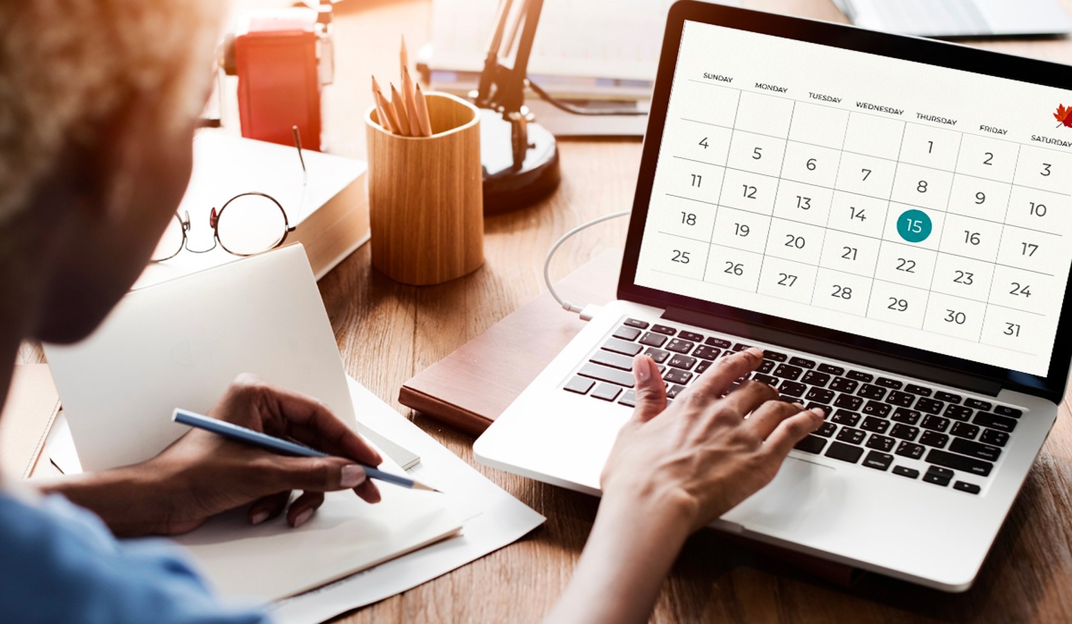 A person writing in a notebook with a pencil in front of a laptop displaying a calendar, highlighting the 15th, on a wooden desk with office supplies.