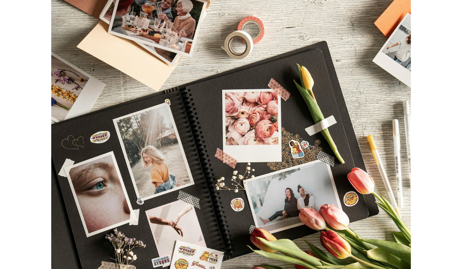 A scrapbook with photos, tulip flowers, stickers, and decorative tape on a light wooden surface, surrounded by pens and washi tapes.