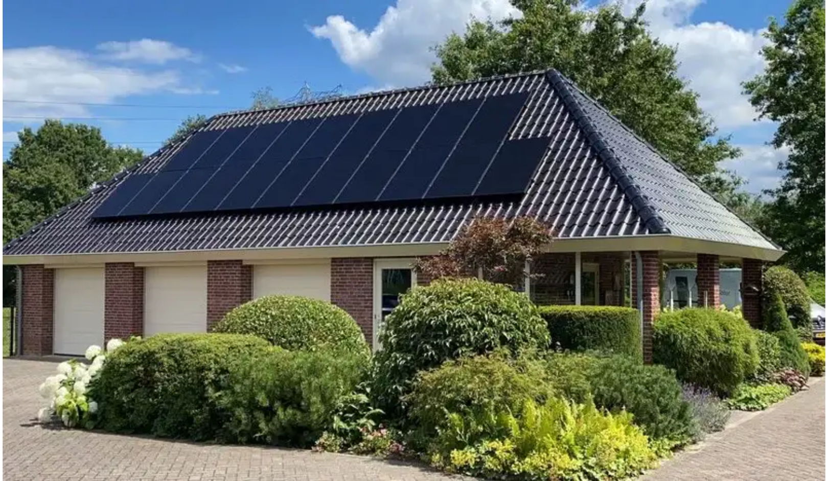 House with a sloped roof featuring solar panels, surrounded by lush green shrubs and trees on a sunny day.
