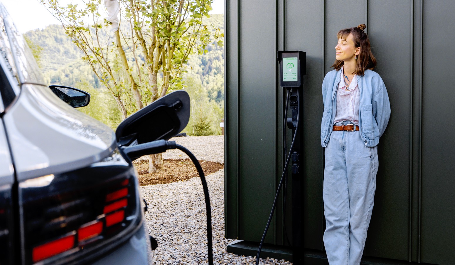 Woman in casual clothes leans against an EV charging station, with an electric car plugged in nearby, set against a backdrop of trees and hills.