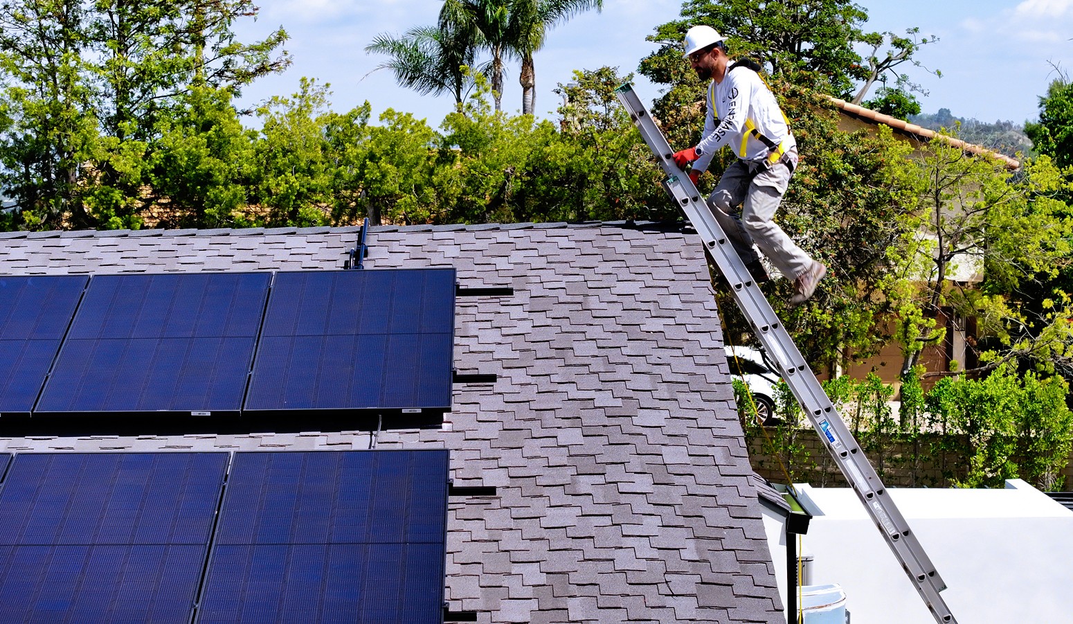 A solar roof contractor in a safety harness climbs a ladder onto a shingled roof with installed solar panels, surrounded by trees.
