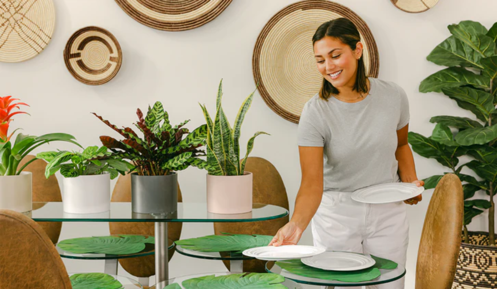 A woman sets a glass table with white plates in a room decorated with potted plants and woven wall art.