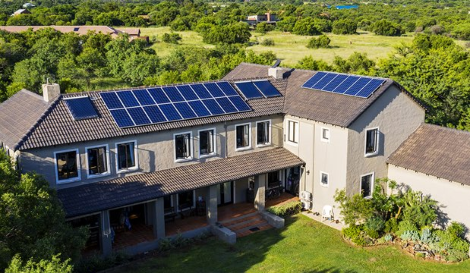 A large house with solar panels on the roof, surrounded by lush greenery and gardens.