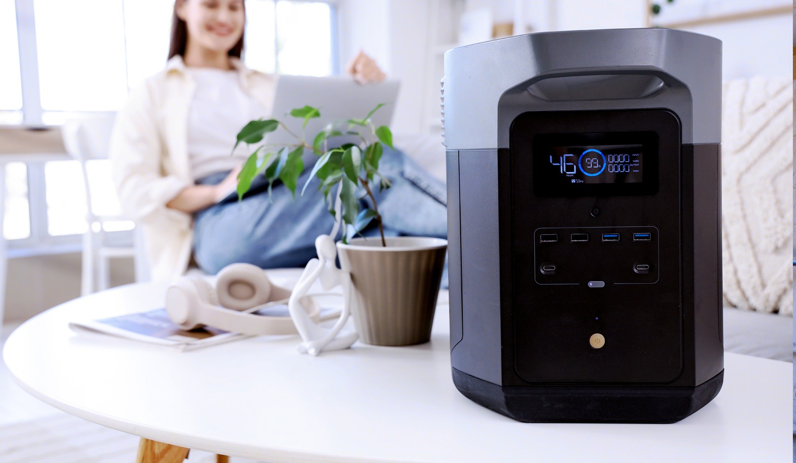 A portable power station on a table, with a potted plant and headphones nearby; a person using a laptop is blurred in the background.