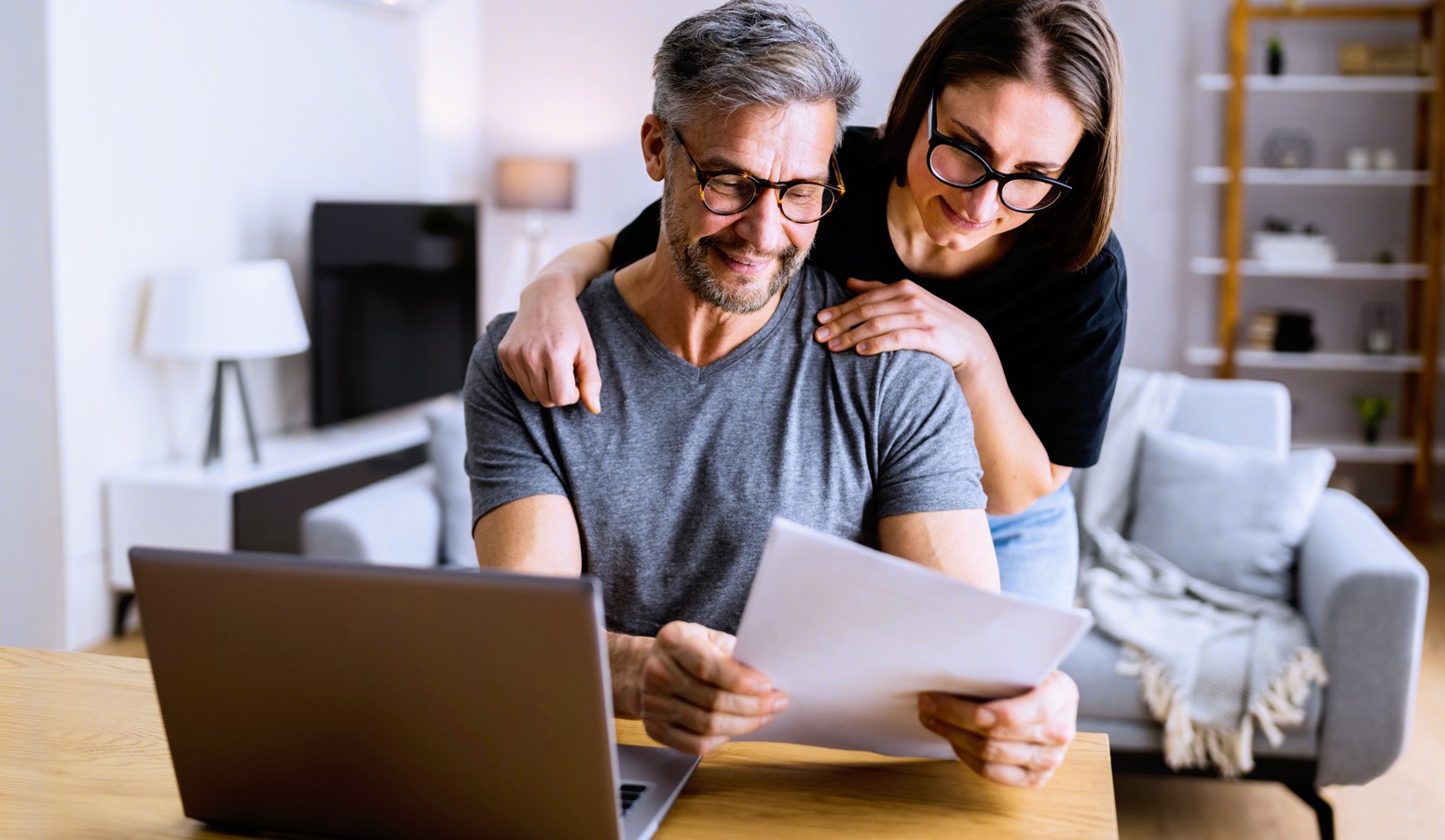 A man and woman smiling while looking at papers in front of a laptop, sitting in a cozy living room with modern decor.