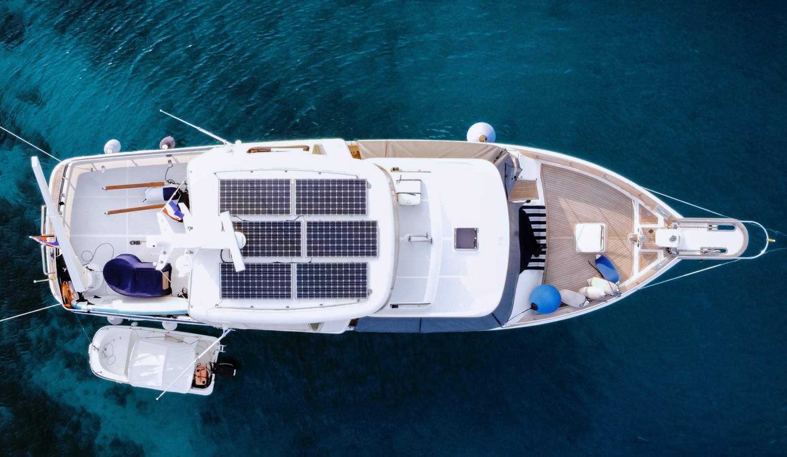Aerial view of a yacht with solar panels on the roof, anchored in clear blue water, with a small boat attached to its side.