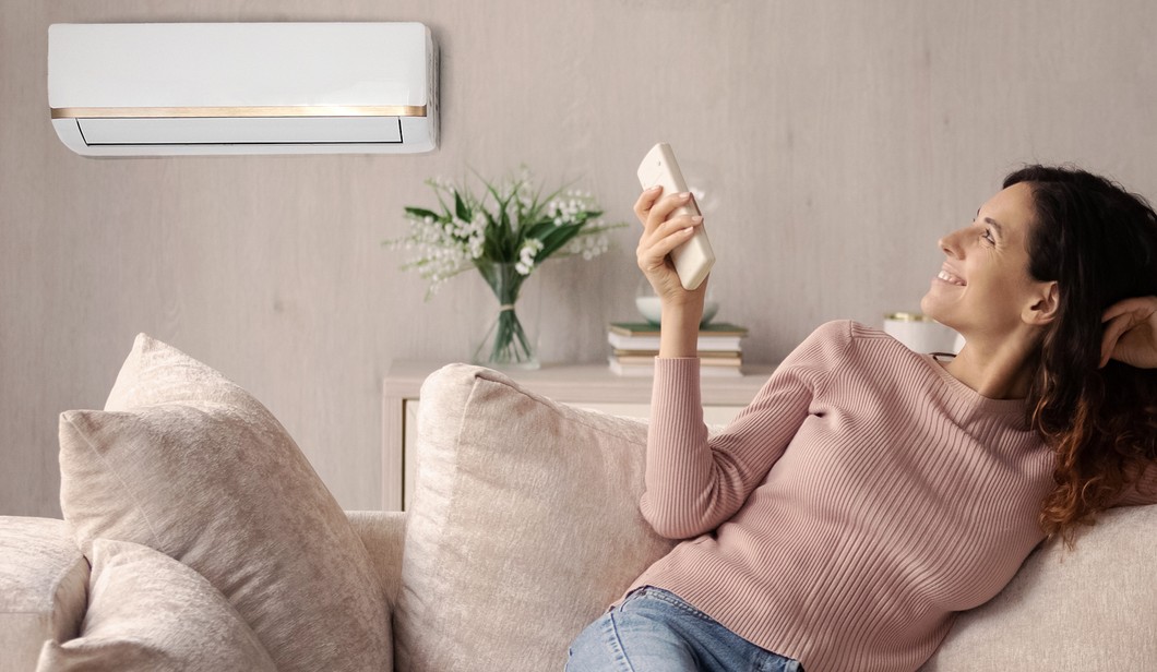 A woman sitting on a beige couch uses a remote to adjust a wall-mounted air conditioner. A vase with flowers is on the table behind her.