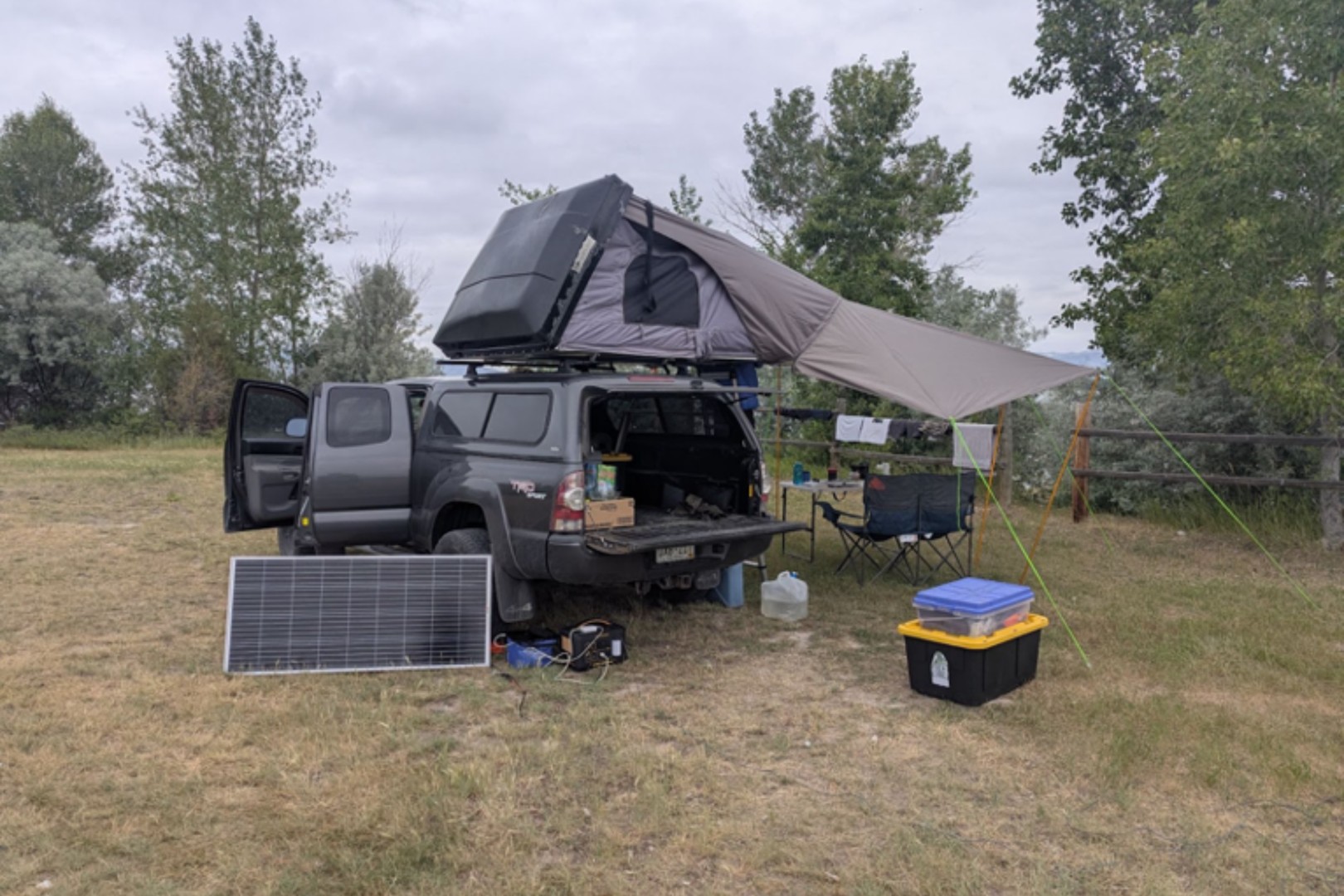 A truck with a rooftop tent, awning, and open tailgate is set up for camping. A solar panel, chair, and storage bin are nearby on grassy ground.