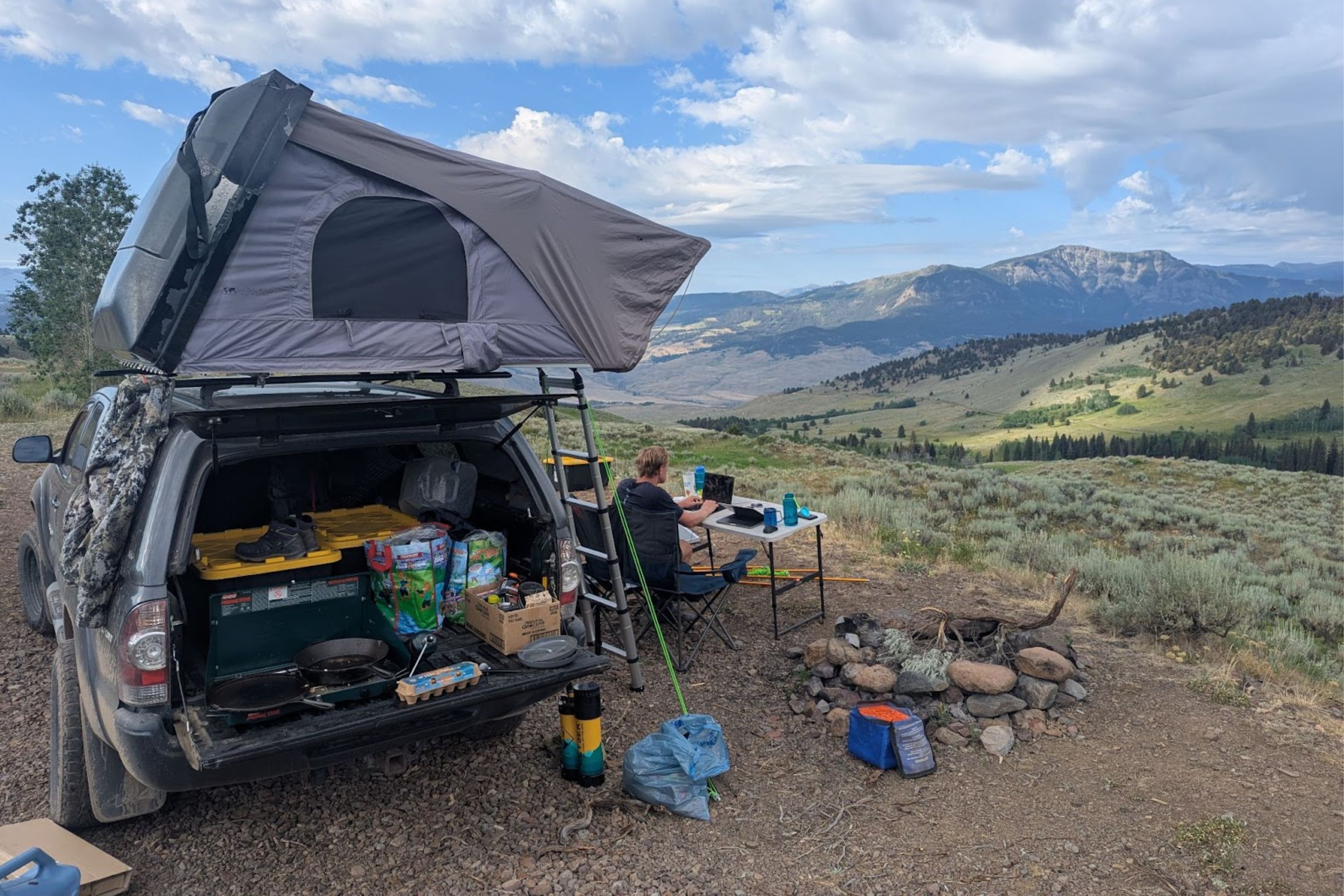 A person camps on a hillside with a roof tent on a vehicle, cooking gear, and a scenic mountain view under a cloudy sky.