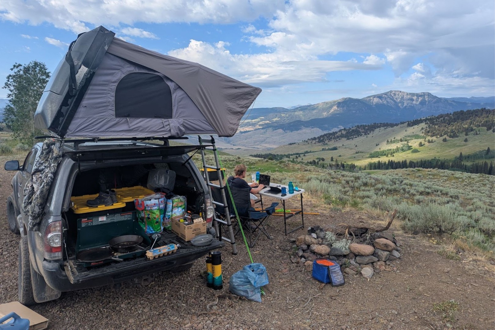 A person camps on a hillside with a roof tent on a vehicle, cooking gear, and a scenic mountain view under a cloudy sky.