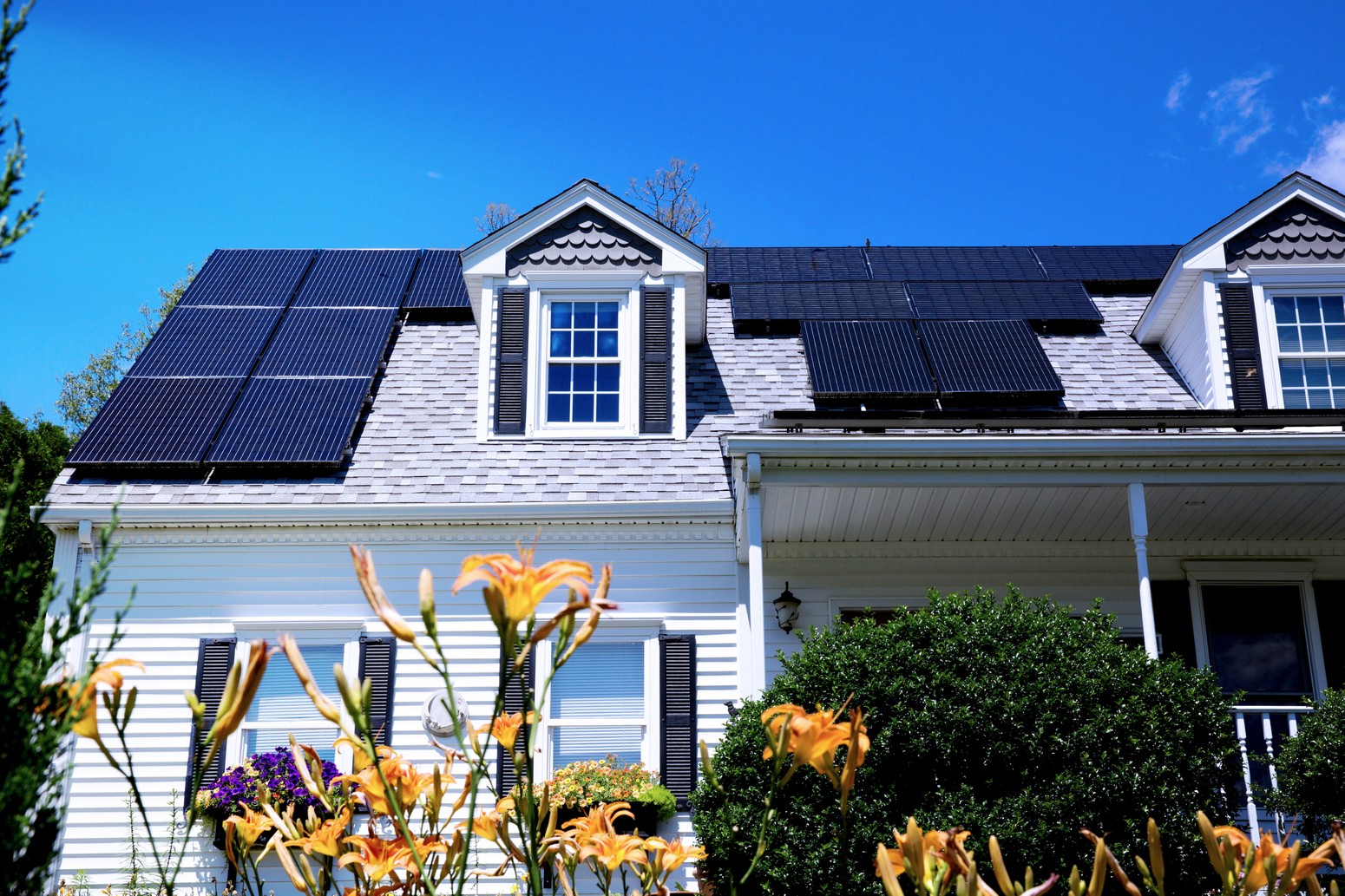 A white house with solar panels on the roof and tiger lily flowers on the front lawn.