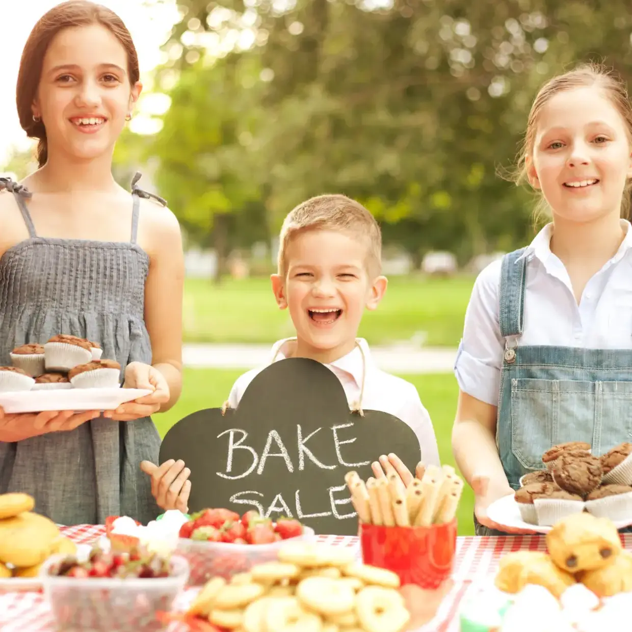 Children smiling at an outdoor bake sale table with treats and a chalkboard sign, representing a Best Fundraiser for schools option and community groups.