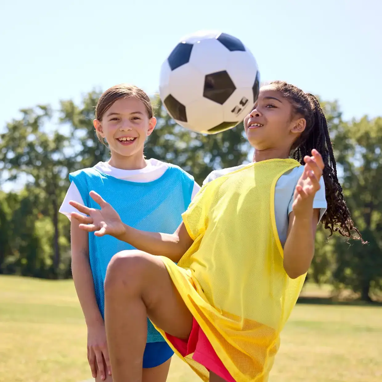 Two kids in sports jerseys play soccer, one kicking the ball mid-air, smiling on a sunny day in the park, trees in the background.