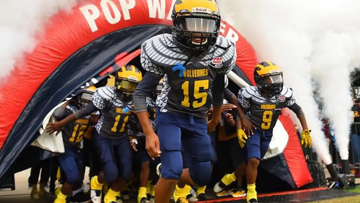 Young football players in blue and yellow uniforms run out of an inflatable tunnel with smoke, preparing for a game.