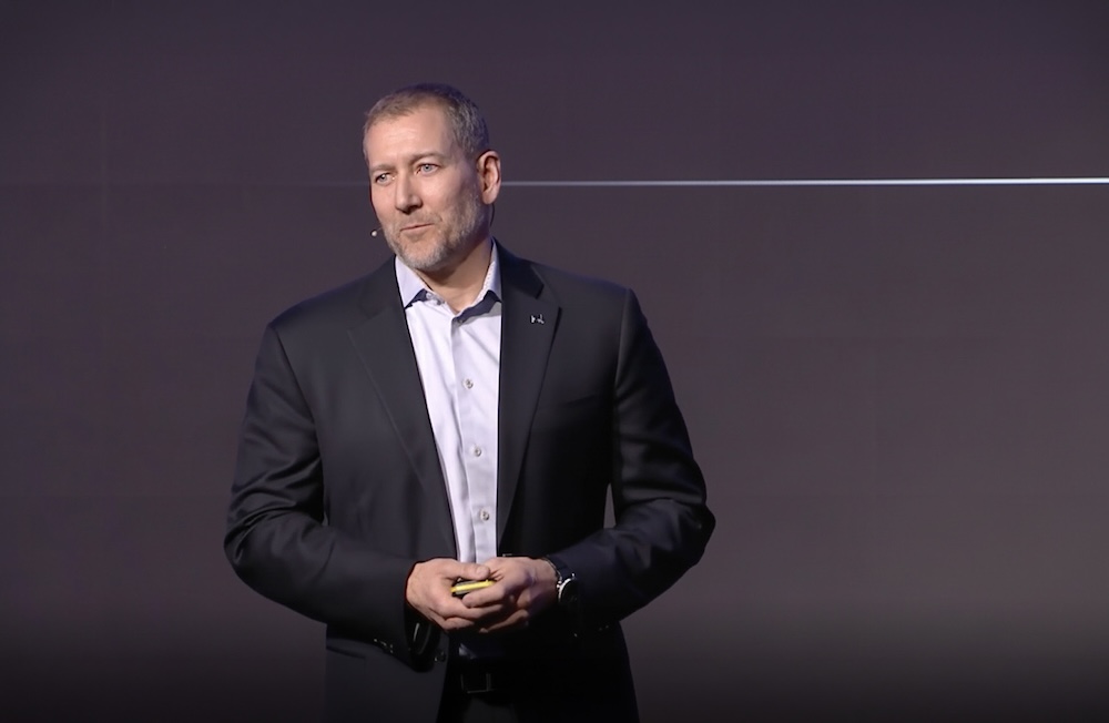 Dan Higgins in a suit giving a presentation on stage, holding a clicker, with a dark background.