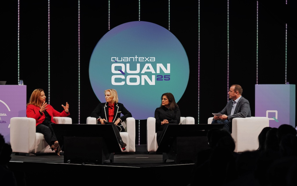 Four panelists seated on stage at QuanCon 25, engaged in discussion, with a large event logo displayed behind them.