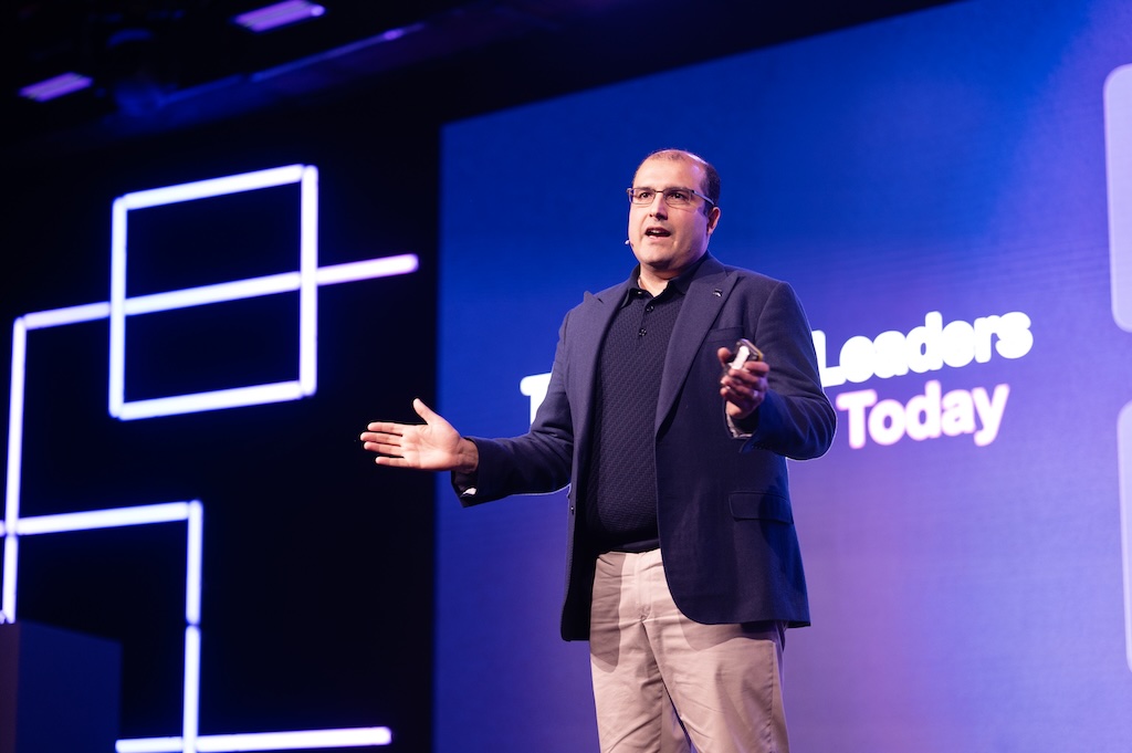 Vishal Marria in a blazer and khakis speaks energetically on stage with a screen displaying partial text and geometric designs in the background.