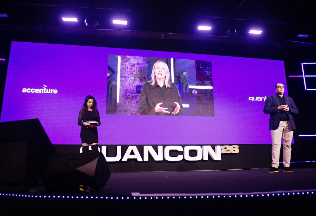 A man and woman stand on stage at Quancon 26, with a large screen showing a woman speaking, logos of Accenture and Quanter visible.