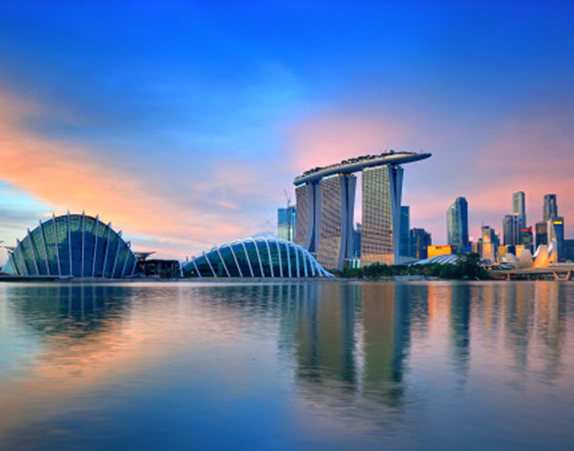 City skyline at sunrise with modern architecture, including curved structures and high-rise buildings, reflected in calm water, under a colorful sky.