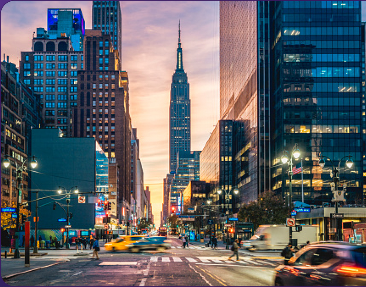 City street scene at dusk with busy traffic, pedestrians, and tall buildings; a prominent skyscraper is visible in the background against a colorful sky.