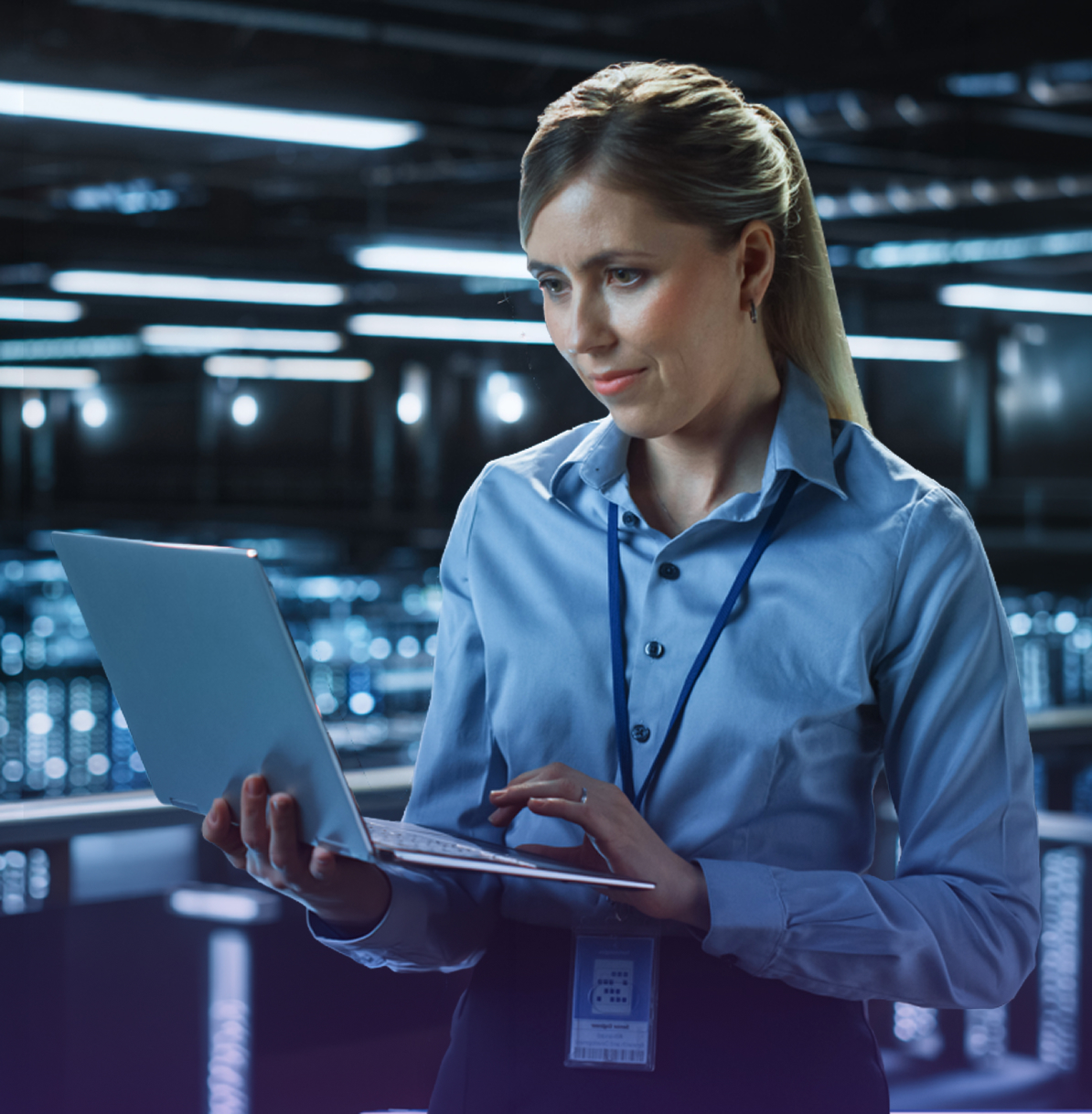 A woman in a blue shirt works on a laptop in a modern data center, surrounded by glowing equipment and dark ambient lighting.