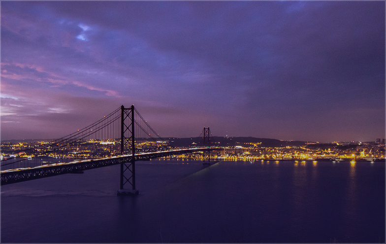 A suspension bridge spans a river at dusk, with city lights glowing beneath a cloudy purple and blue sky.