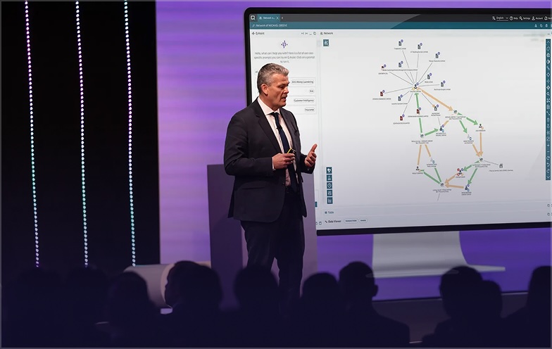 A man in a suit presents data on a large screen displaying a flowchart, in front of an audience.