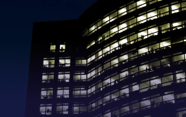 A tall building at night with illuminated windows revealing silhouettes of people working inside.