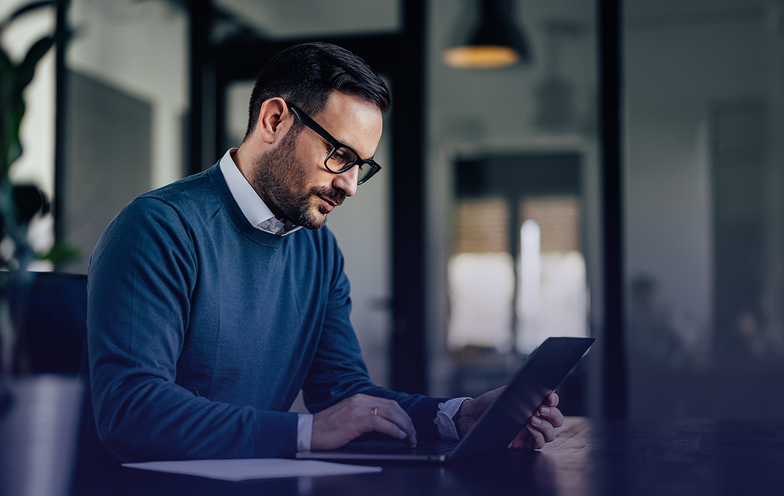 Man in glasses and blue sweater working on a laptop at a desk in a dimly lit office.