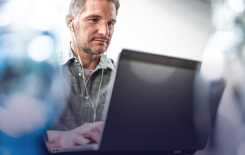 Man in a gray shirt using a laptop, wearing earphones, focused on his work. Blurred blue elements in the foreground.