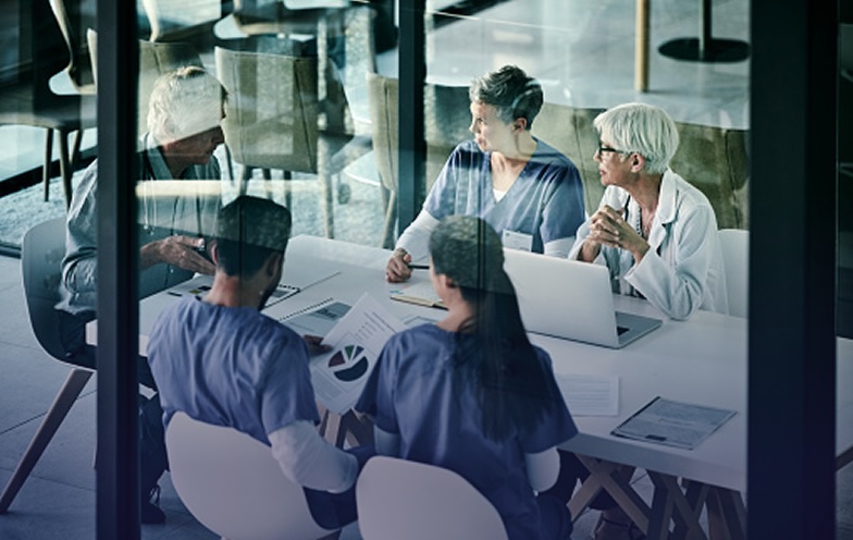 A group of medical professionals in scrubs and lab coats having a meeting around a table with laptops and documents.