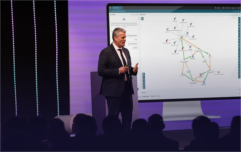 A man in a suit presents on stage with a large screen displaying a network diagram, in front of an audience.