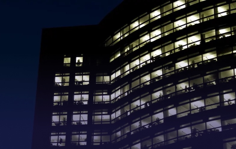 Office building at night with illuminated windows, revealing silhouettes of people working inside.