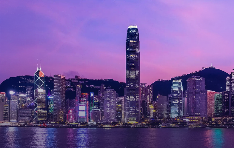 Skyline of a city with illuminated skyscrapers against a purple and pink sunset sky, reflecting on a calm body of water in the foreground.